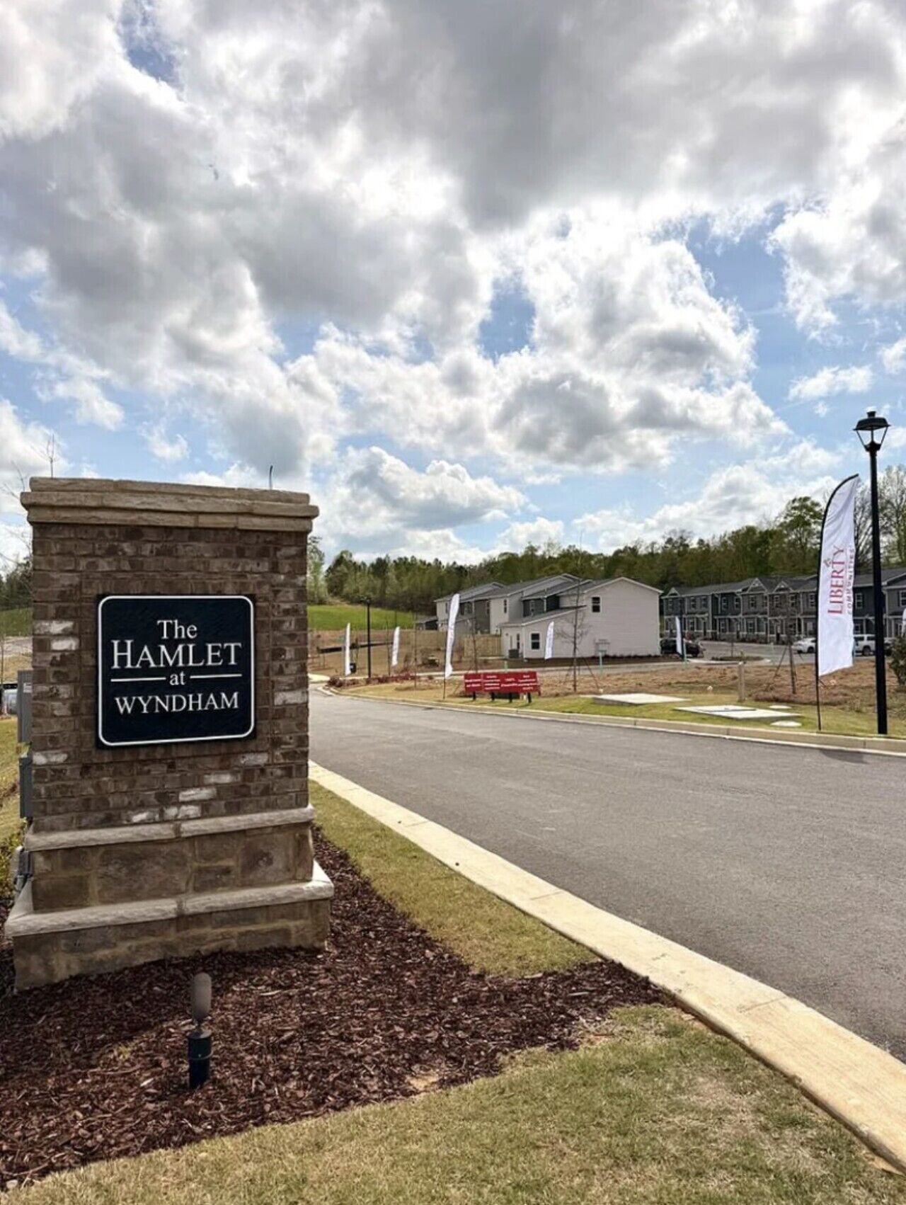 A stone sign with 'The Hamlet at Wyndham' text, a paved road, a building in the background, and a partly cloudy sky.