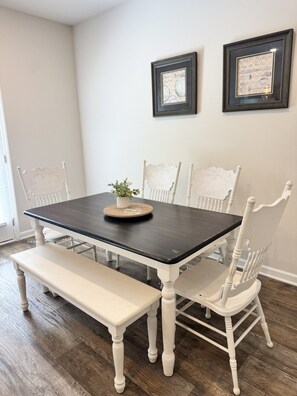A dining table with a dark top, surrounded by white chairs with ornate backs. A small potted plant is on the table, and there are two framed pictures on the wall above.