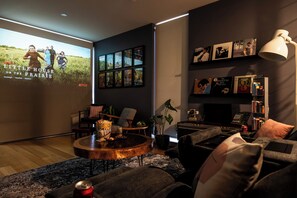 A modern living room with a large screen displaying a movie poster, comfortable seating, a bookshelf, and a coffee table. - Spacious Vibey Retreat with a Movie Projector, Electric Recliner and & Balcony (Melbourne)