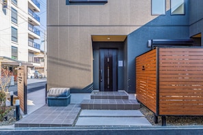 A modern building with a flat facade, a dark entrance door, and a small outdoor seating area with a trash bin. - Apartment Hotel 11 Villa Tengachaya (Osaka-shi)