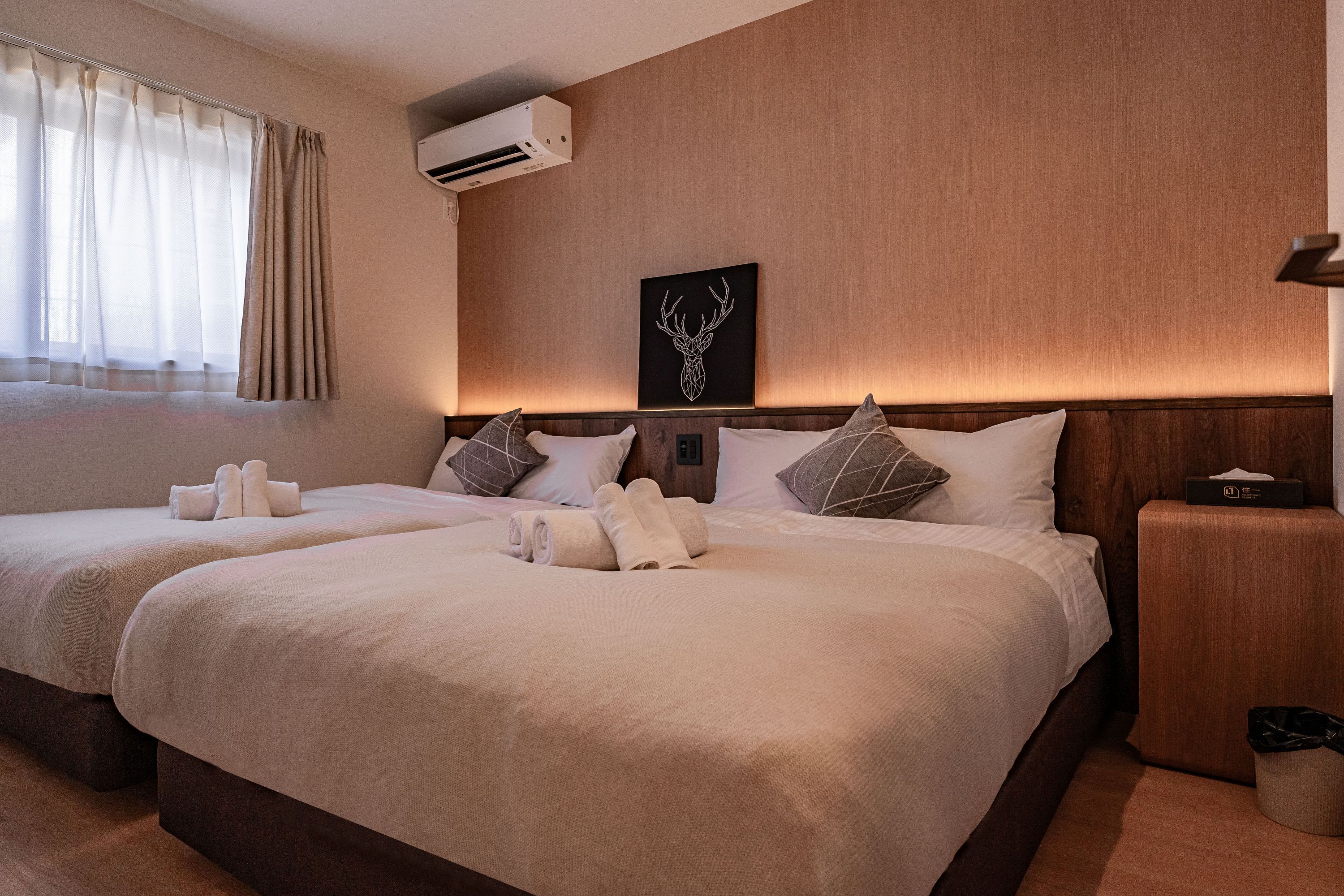 Camera d'albergo con due letti, condizionatore d'aria a parete, un quadro con testa di cervo incorniciata e un comodino con un portaserviti.