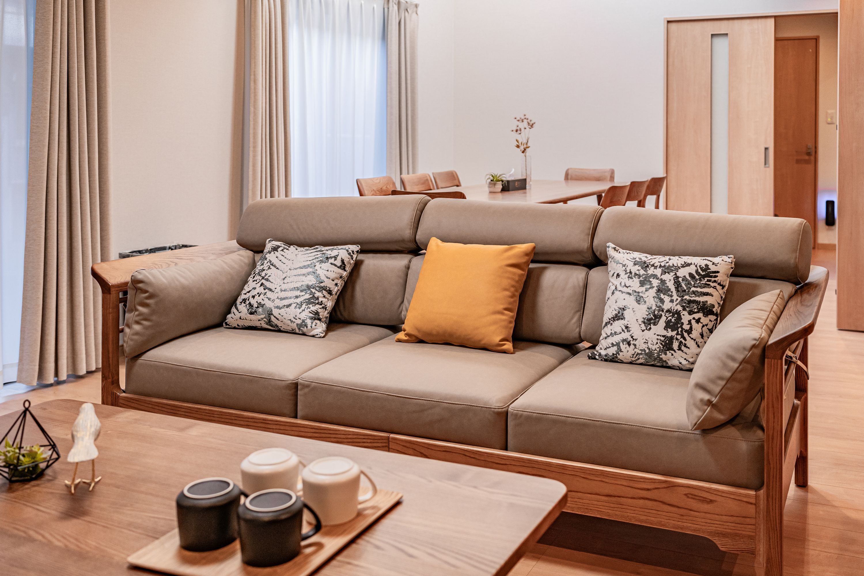 A modern living room with a beige sofa, patterned cushions, a wooden coffee table with mugs, and a dining area in the background.