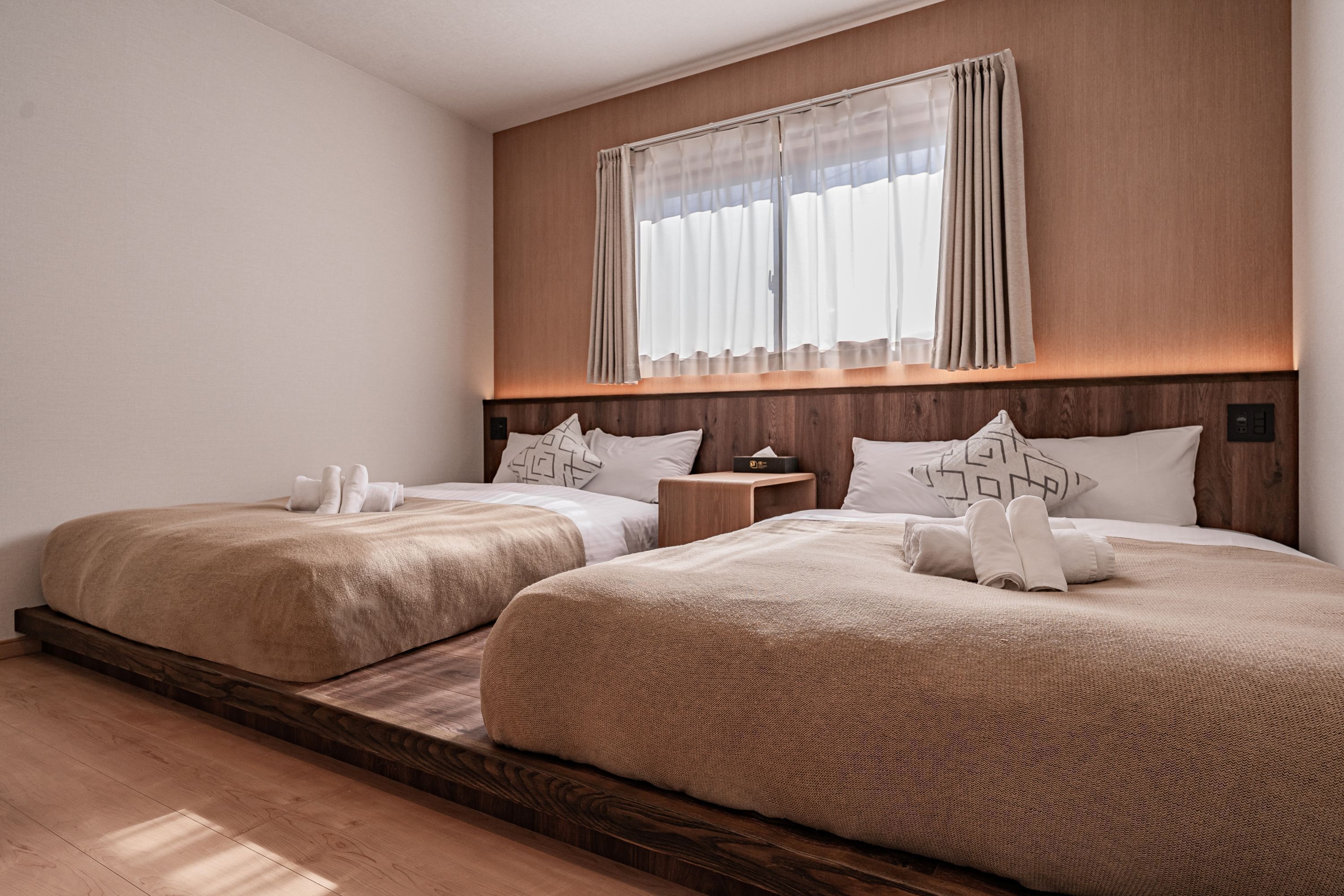 Habitación de hotel con dos camas, cabecero de madera, mesita de noche y ventana con cortinas.