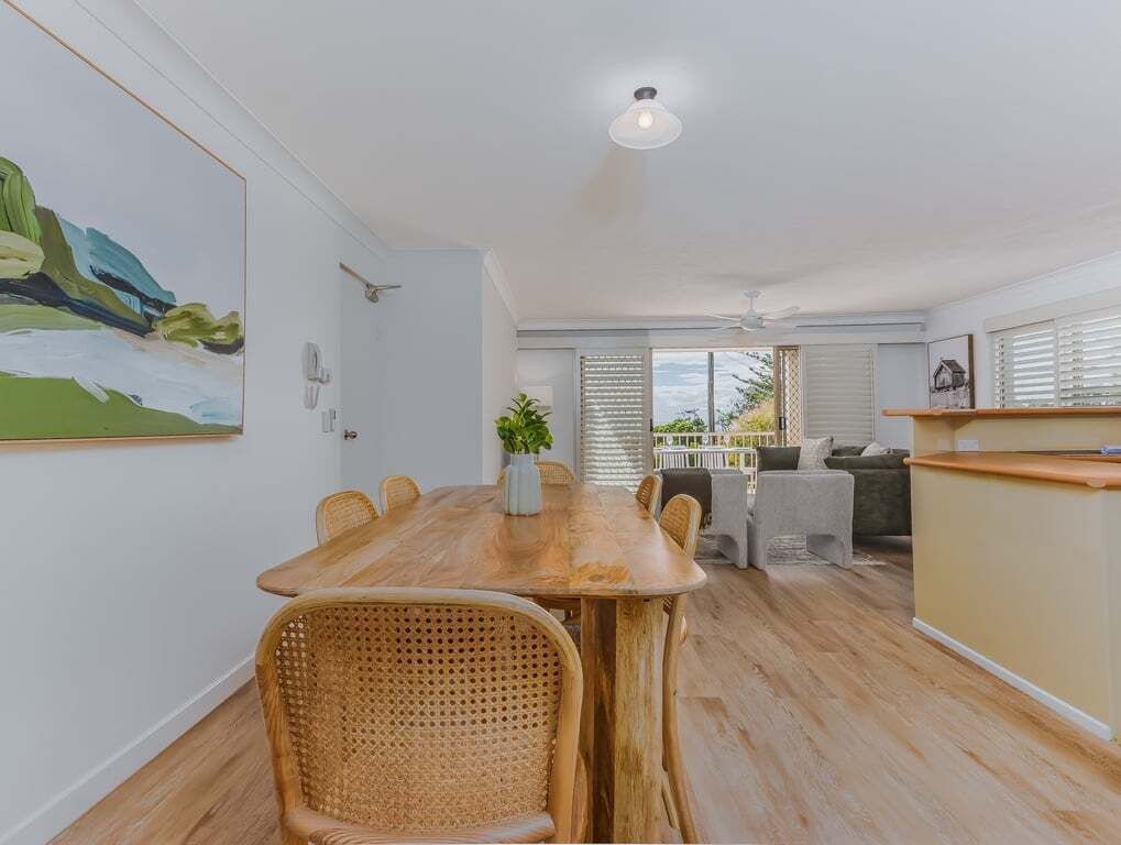 A dining area with a wooden table and wicker chairs, a kitchen area with a counter, and a view of the outdoors through a window.
