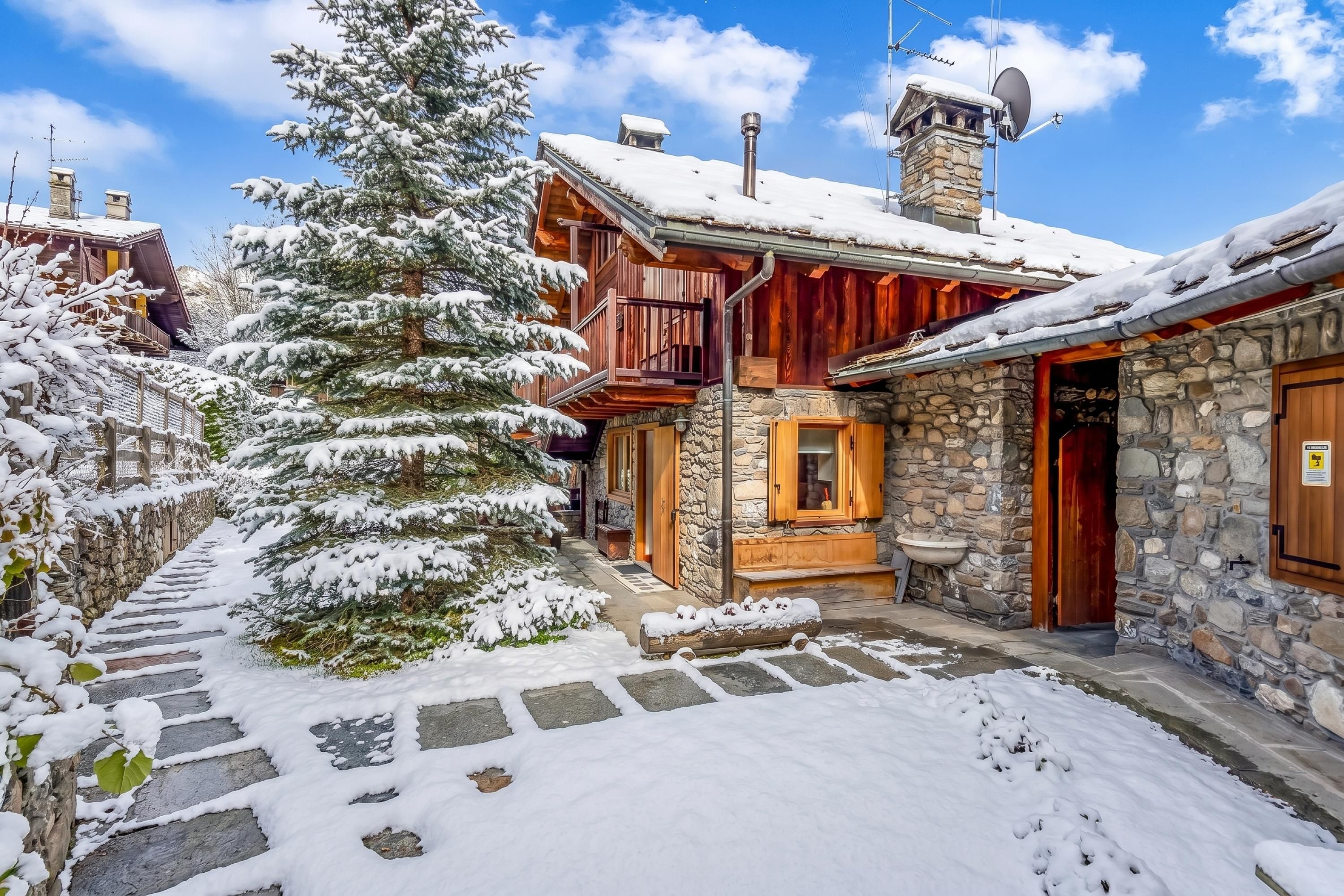 Un edificio de madera cubierto de nieve con muros de piedra, una escalera de piedra y un árbol nevado.