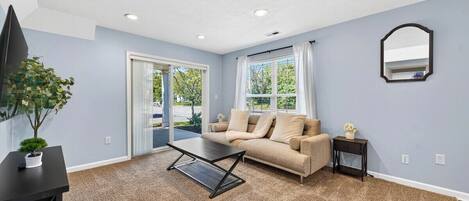 A room with a beige sofa, a coffee table, a small side table with a vase, a potted plant, and a mirror on the wall.