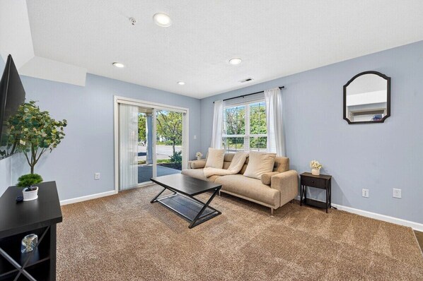 Une pièce avec un canapé beige, une table basse, une petite table d’appoint avec un vase, une plante en pot et un miroir au mur.