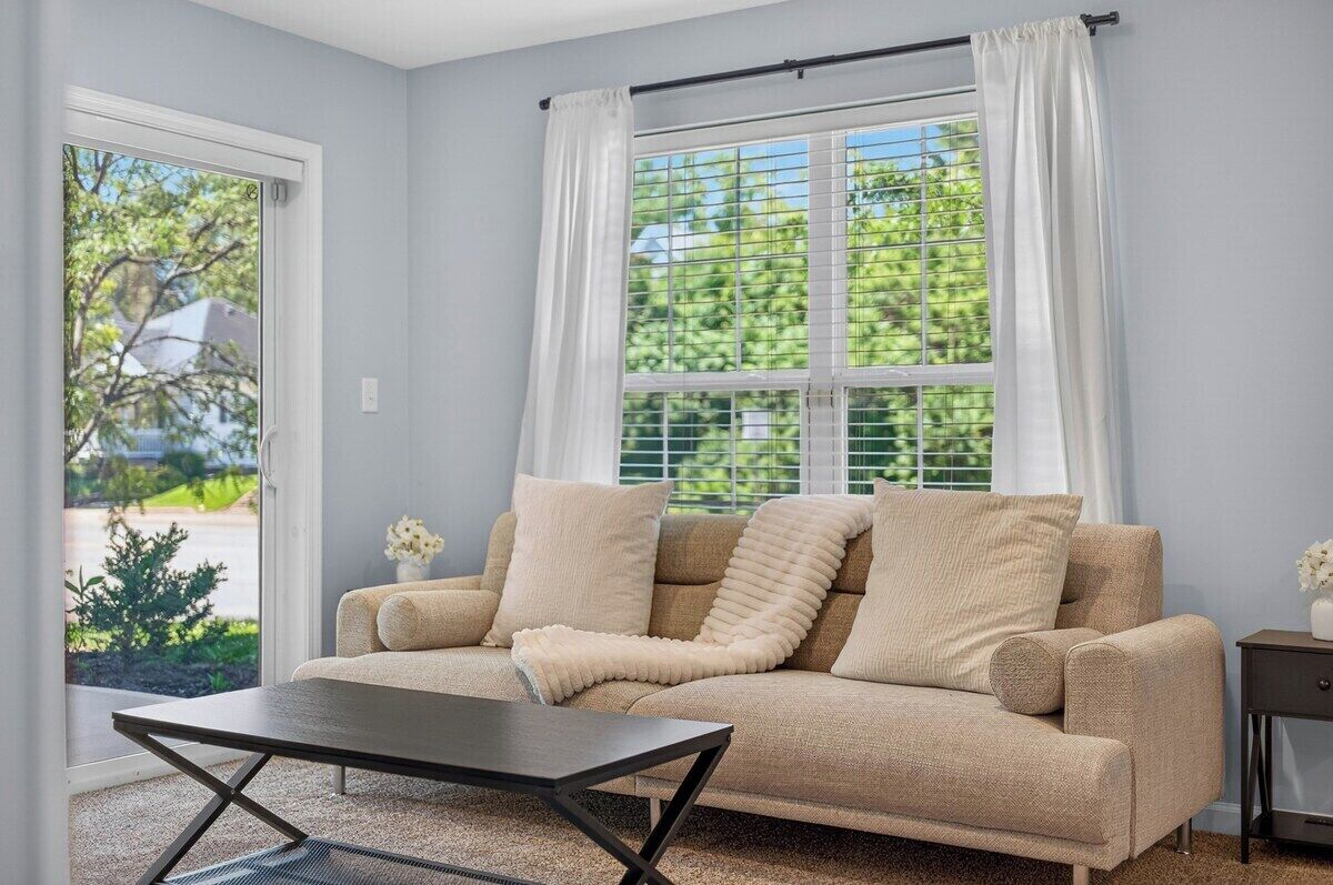 A living room with a beige sofa, a coffee table, and a window with white curtains.