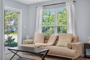 A living room with a beige sofa, a coffee table, and a window with white curtains.