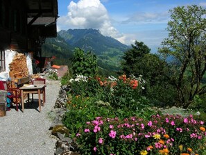Um edifício rústico com uma mesa e cadeiras de madeira à mostra, rodeado por flores coloridas e vista para montanhas.