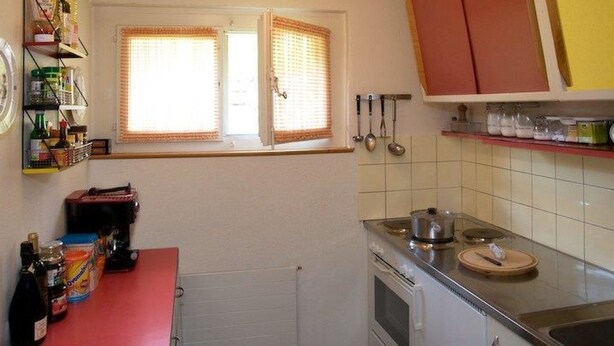 Een keuken met een rode aanrecht, een fornuis en een raam met zonwering.