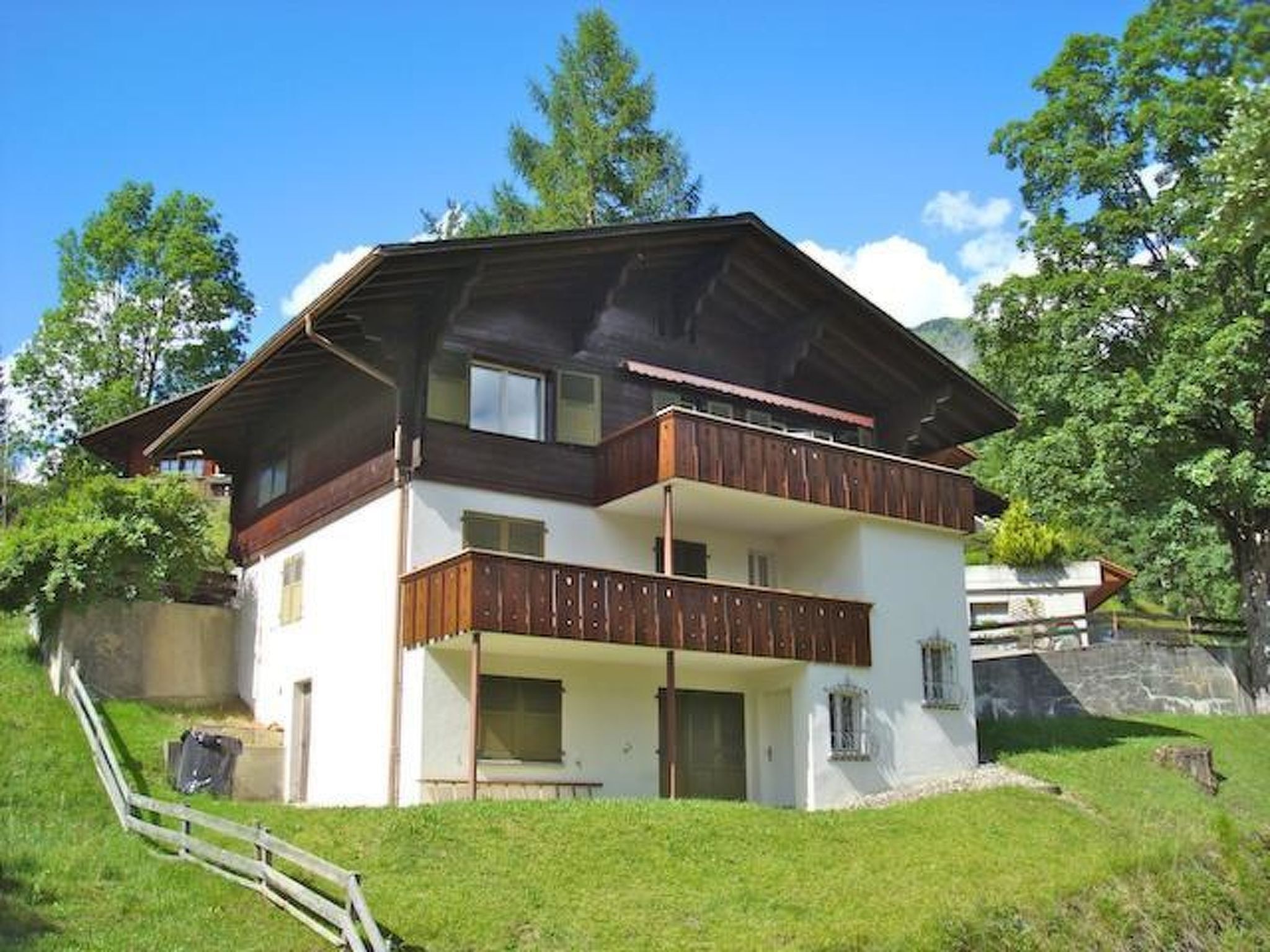 Une maison à deux étages avec un balcon à l’étage supérieur, un porche à l’avant et un mur de pierre en arrière-plan.