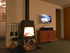 A living room with a wood-burning stove, a television mounted on the wall, and a wooden cabinet. - Pony 5-Bettwohnung Appartement/Fewo, Dusche oder Bad, WC, 2 Schlafräume by Interhome (Lenk)