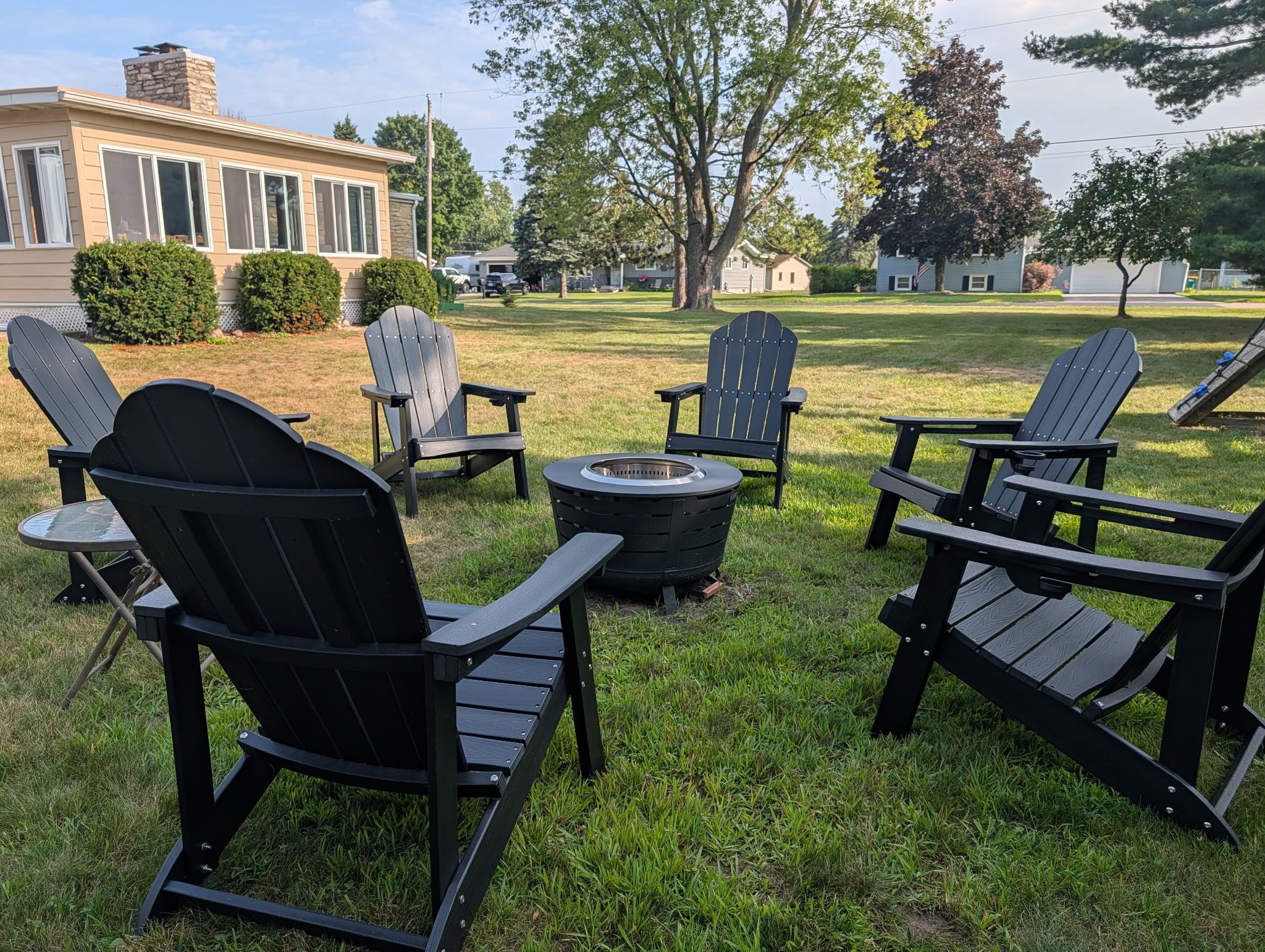 Un patio trasero con mobiliario exterior, incluyendo sillas Adirondack y una mesa redonda, un brasero y un césped bien cuidado.