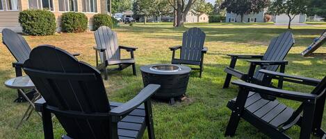 Un patio trasero con mobiliario exterior, incluyendo sillas Adirondack y una mesa redonda, un brasero y un césped bien cuidado.