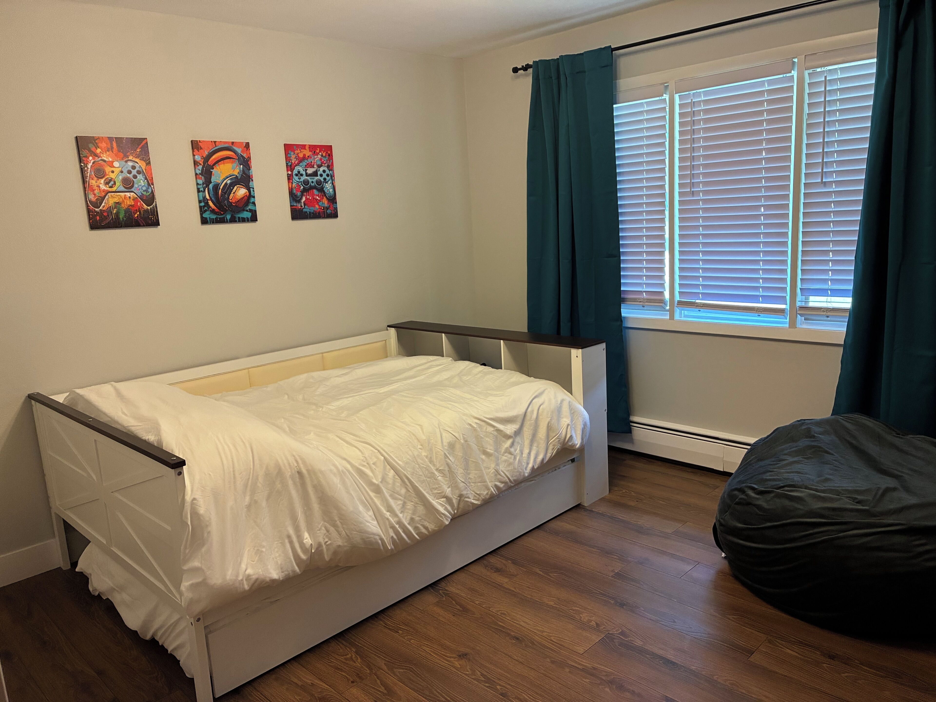 A single-bed room with a wooden floor, a window with blinds, and three wall-mounted paintings.