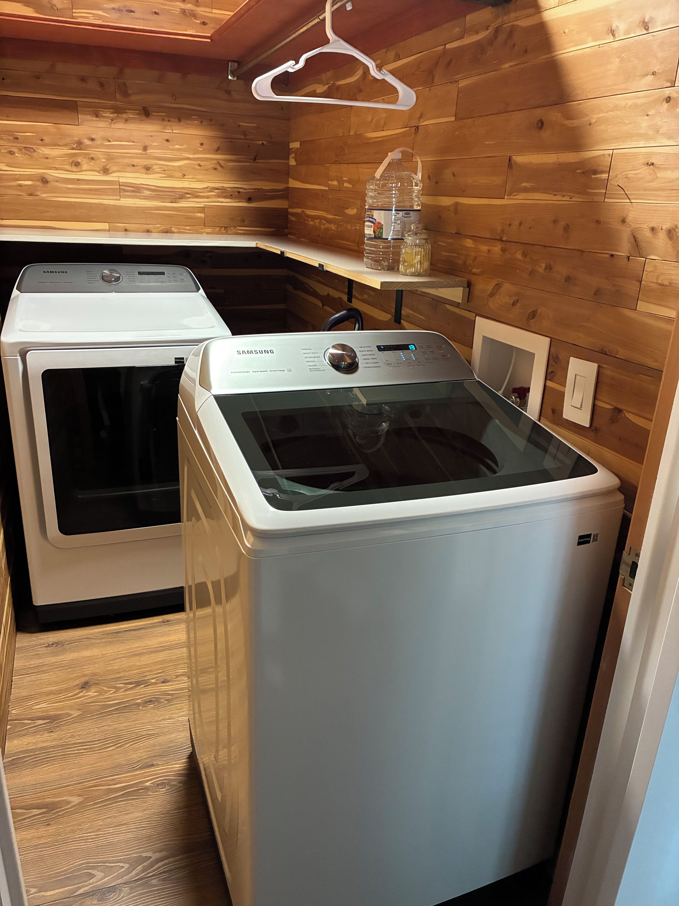 A laundry room with two white Samsung washing machines, wooden walls, and a hanging clothes rack.