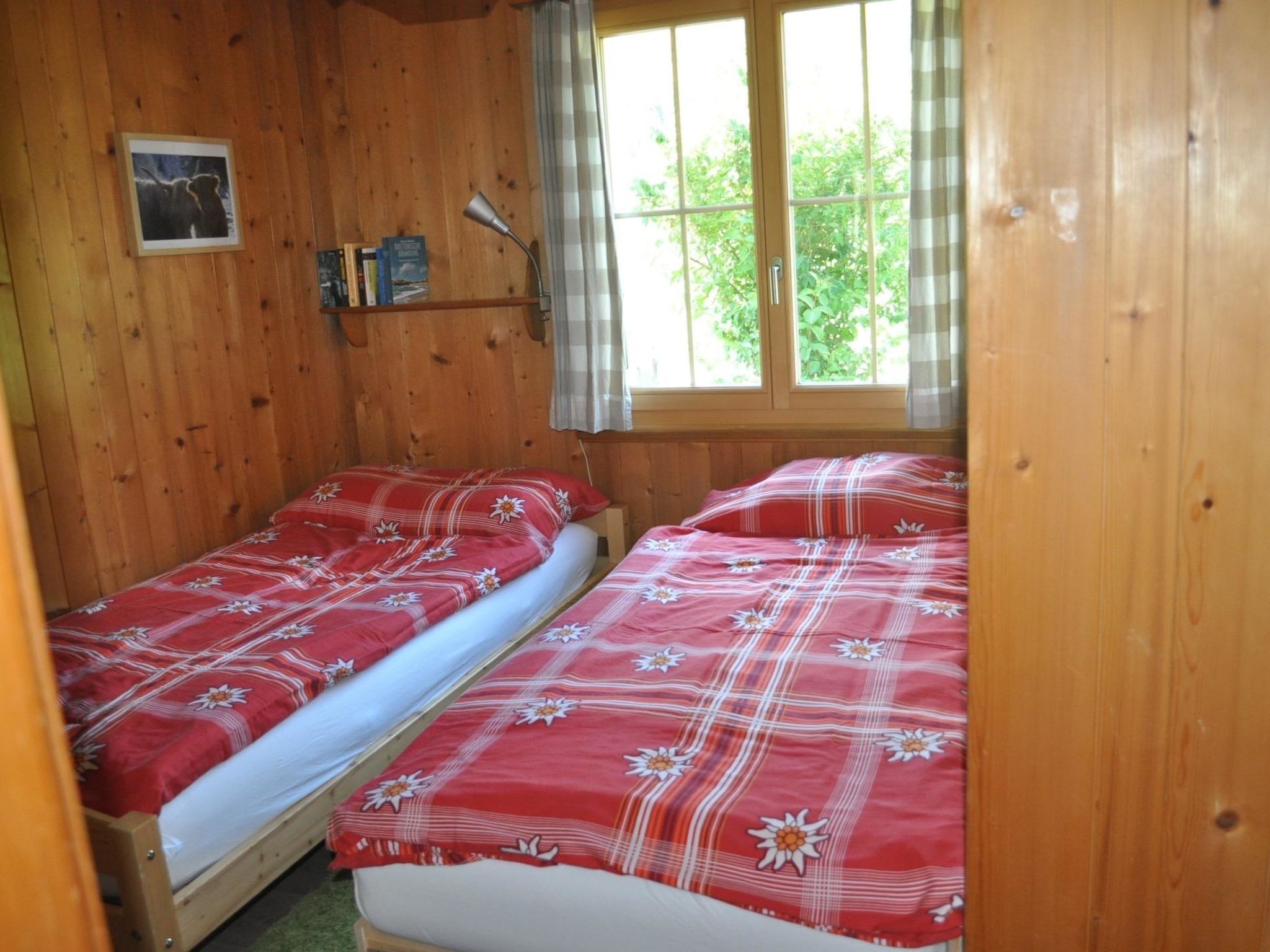 Une chambre dans un chalet en bois, avec deux lits, une fenêtre avec des rideaux et une étagère remplie de livres.