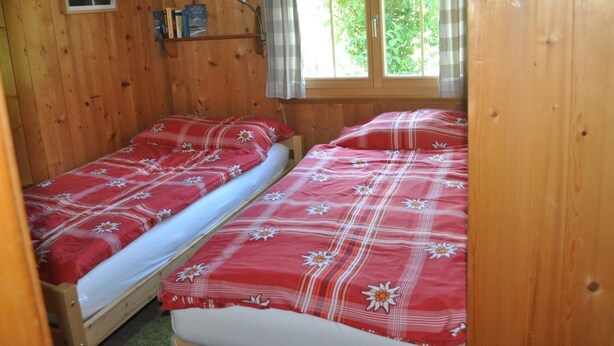 A wooden cabin room with two beds, a window with curtains, and a shelf with books. - Andrea Appartement/Fewo, Dusche oder Bad, WC, 3 Schlafräume 4-Zimmerhaus by Interhome (Lenk)