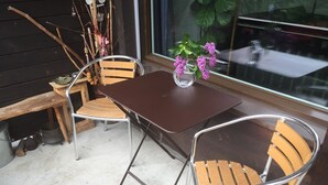 Un pati amb dues cadires de metall i una taula, un vas amb flors i una finestra amb vista a vegetació.