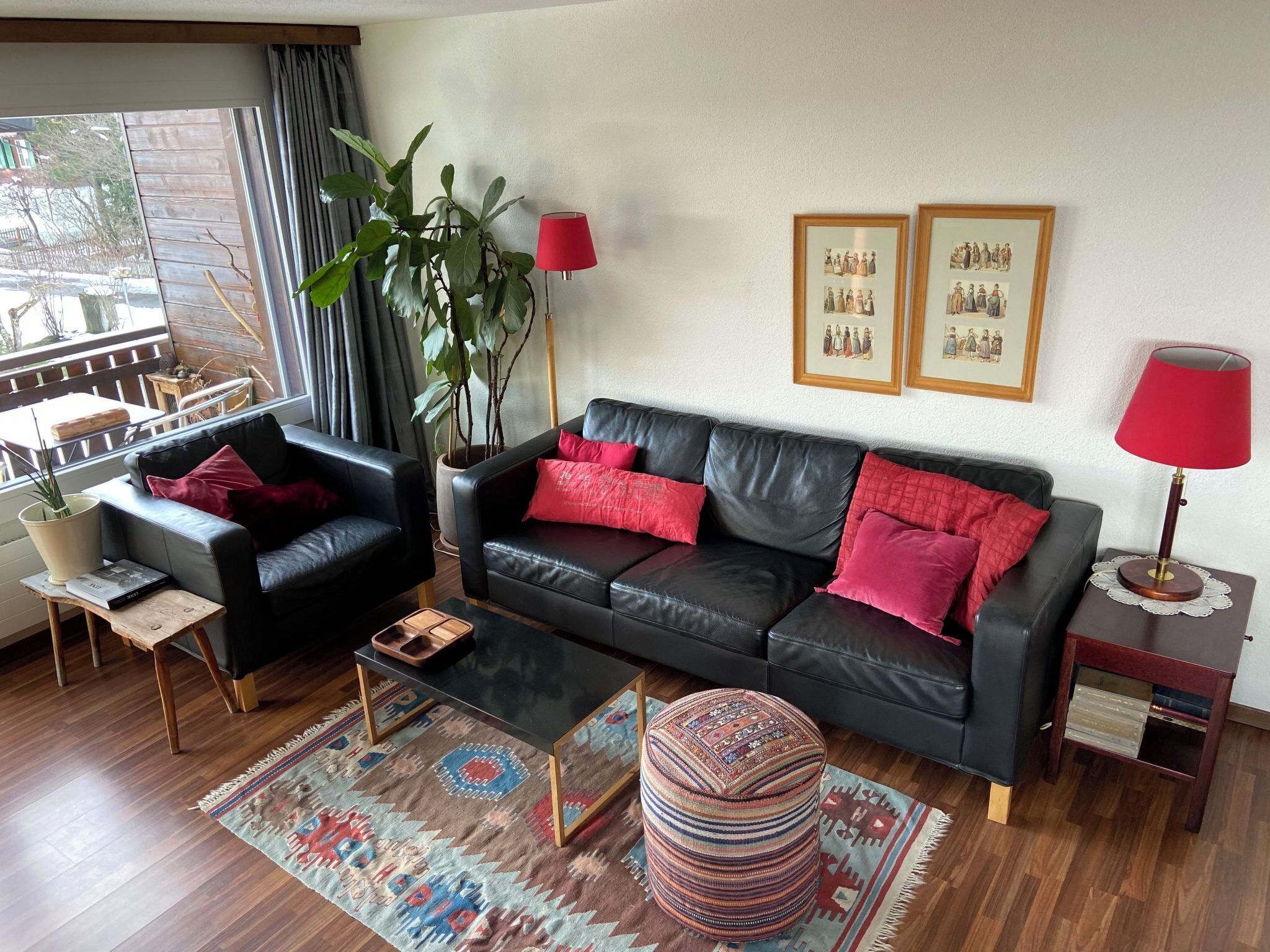 A living room with a black leather sofa, red cushions, a wooden coffee table, a patterned rug, a potted plant, and two framed pictures on the wall.