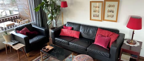Ett vardagsrum med en svart lädersoffa, röda kuddar, ett träbord, en mönstrad matta, en krukväxt och två inramade tavlor på väggen.