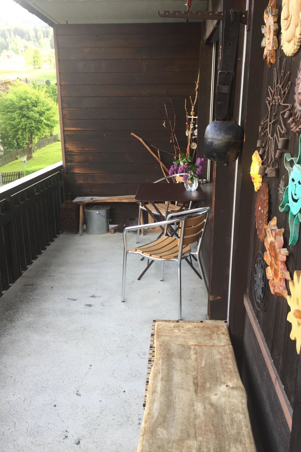 A wooden porch with a metal chair, a small table with a vase, and a bench.