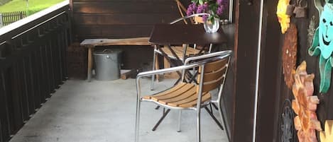 A wooden porch with a metal chair, a small table with a vase, and a bench.
