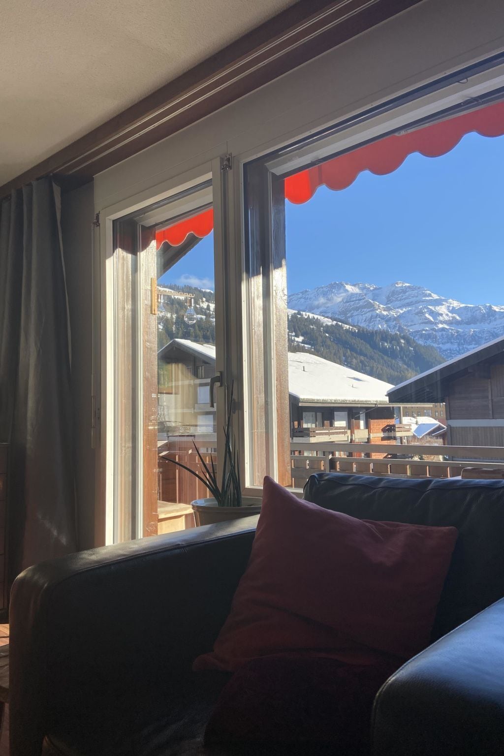 A room with a view of snow-capped mountains, a window with a red frame, and a sofa with a red cushion.