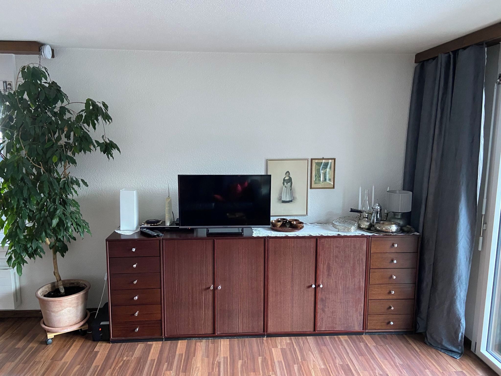 A living room with a flat-screen TV, wooden cabinet, potted plant, and a window with curtains.