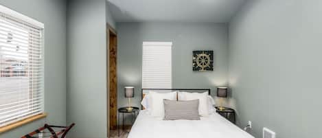 Une chambre à coucher comprenant un lit, deux tables de chevet, une fenêtre avec des stores, une œuvre d’art accrochée au mur et un radiateur.