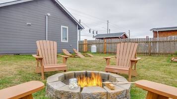 Un patio trasero con un hogar a leña de piedra, dos sillas Adirondack y una mesa de madera.