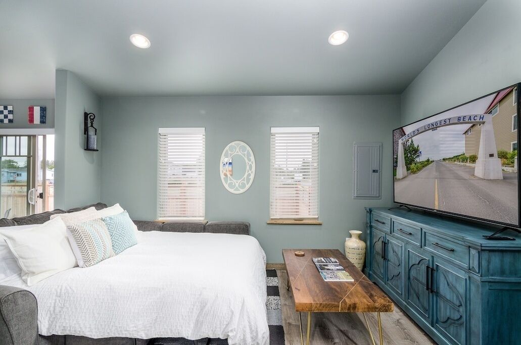 A bedroom with a bed, a television, a dresser, and two windows with blinds.