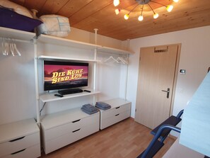 Quarto com televisão sobre uma prateleira branca, uma porta e um lustre.