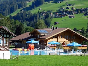 Ein Hotel mit Poolbereich, inklusive Liegestühlen und Sonnenschirmen, vor der Kulisse grüner Hügel und bewaldeter Berge.