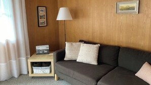 Et værelse med en trævæg, en grå sofa med to puder, en hvid gulvlampe, et lille trænatbord med en lampe, og et billede i ramme på væggen.