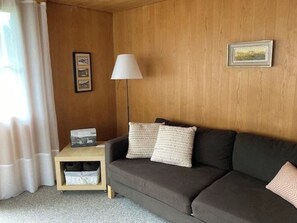 Ein Zimmer mit einer Holzvertäfelung, einem grauen Sofa mit zwei Kissen, einer weißen Stehlampe, einem kleinen Holzhochentisch mit einer Lampe und einem gerahmten Bild an der Wand.