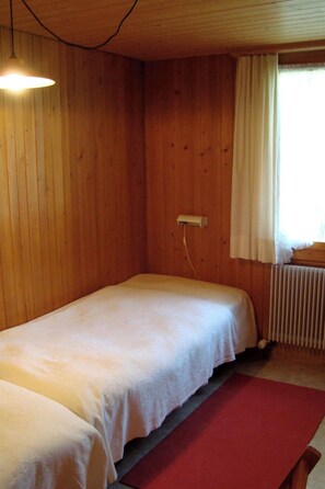 Ein Einzelbett in einem Zimmer mit Holzwänden, einem Heizkörper und einem Fenster mit Vorhängen.