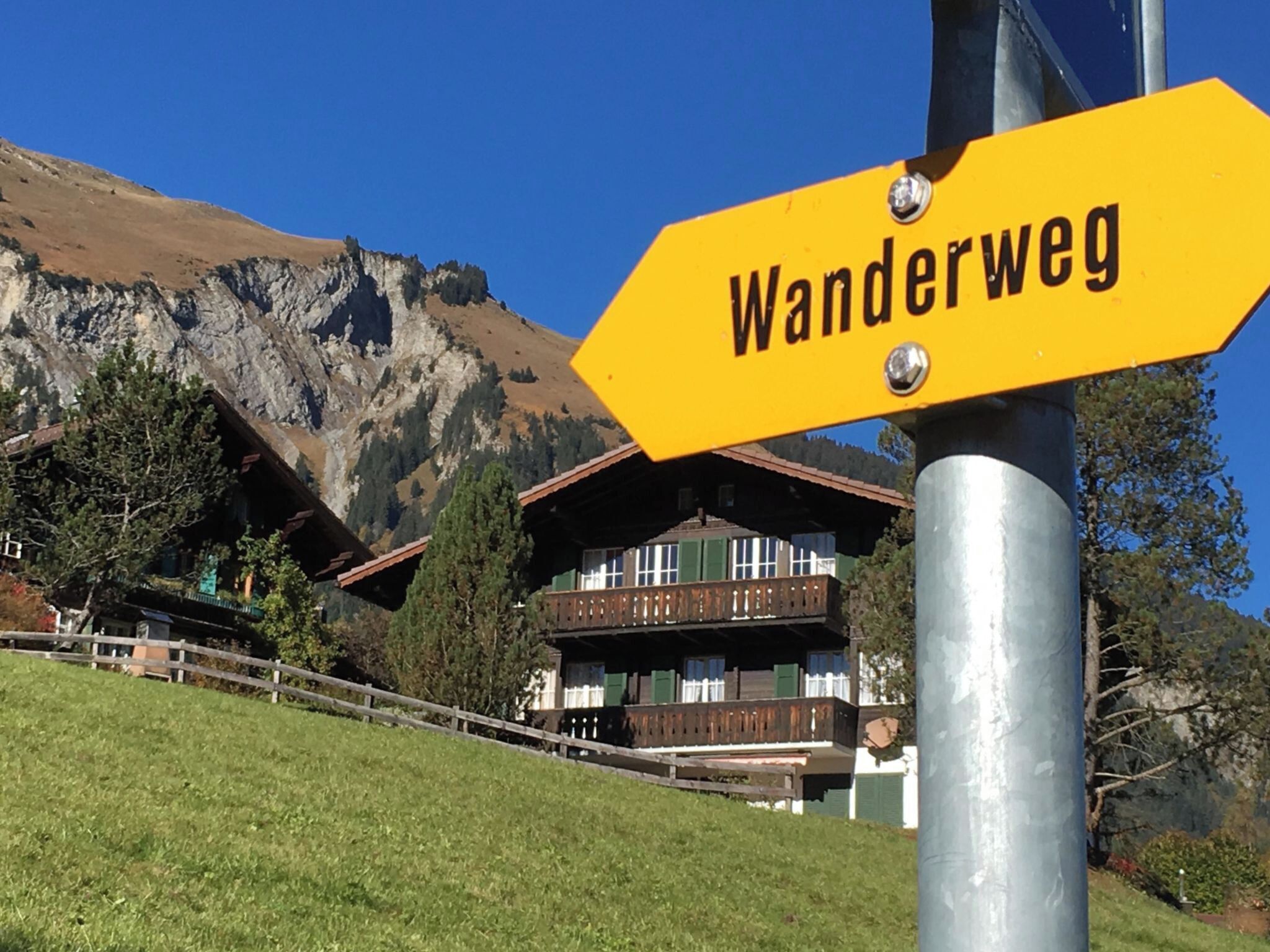 A yellow sign with 'Wanderweg' written on it, a wooden building with a balcony, and a mountain in the background.