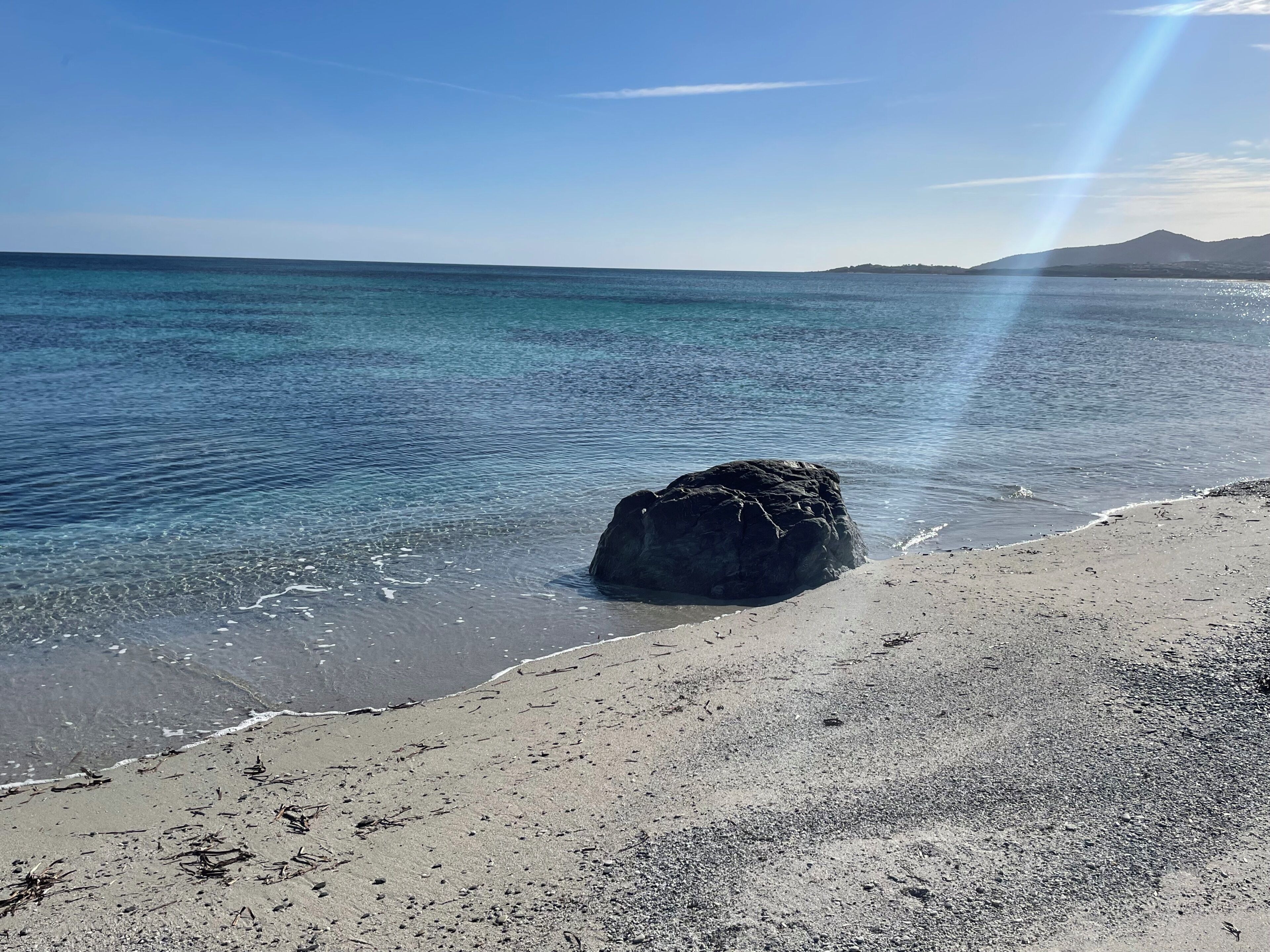 Une plage à l’eau claire, un grand rocher et une chaîne de montagnes lointaine.