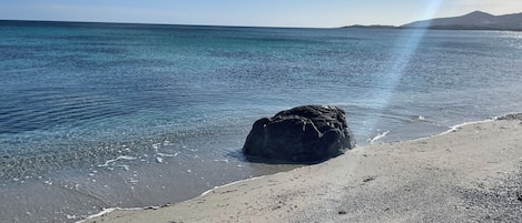 A beach with clear water, a large rock, and a distant mountain range.