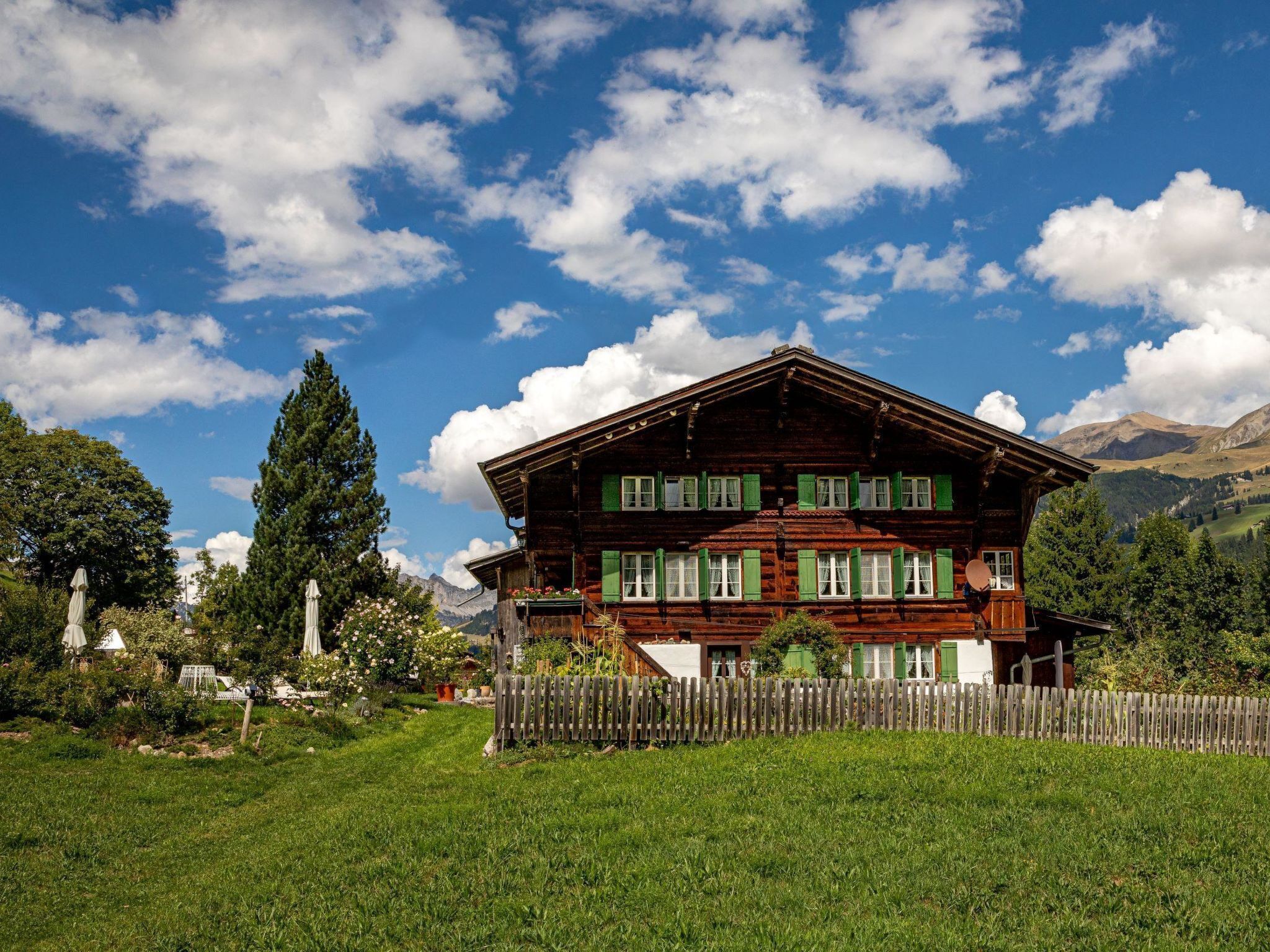 Ein zweistöckiges Holzhaus mit Balkon, umgeben von einem Holzzaun und üppiger Bepflanzung.