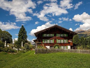 Une maison en bois à deux étages, dotée d’un balcon, entourée d’une clôture en bois et d’une végétation luxuriante.