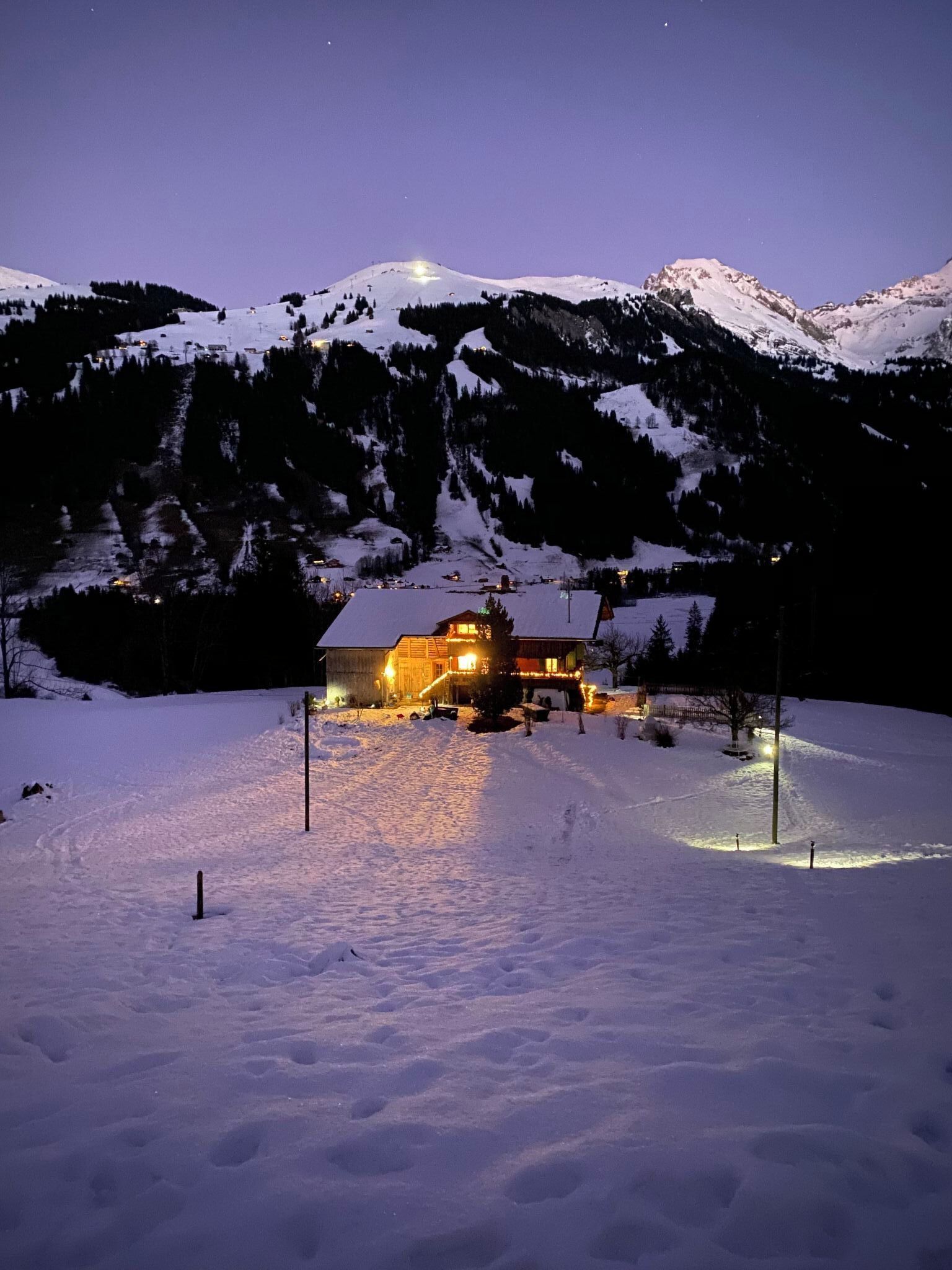 A snow-covered landscape with a lit building in the foreground, surrounded by mountains.
