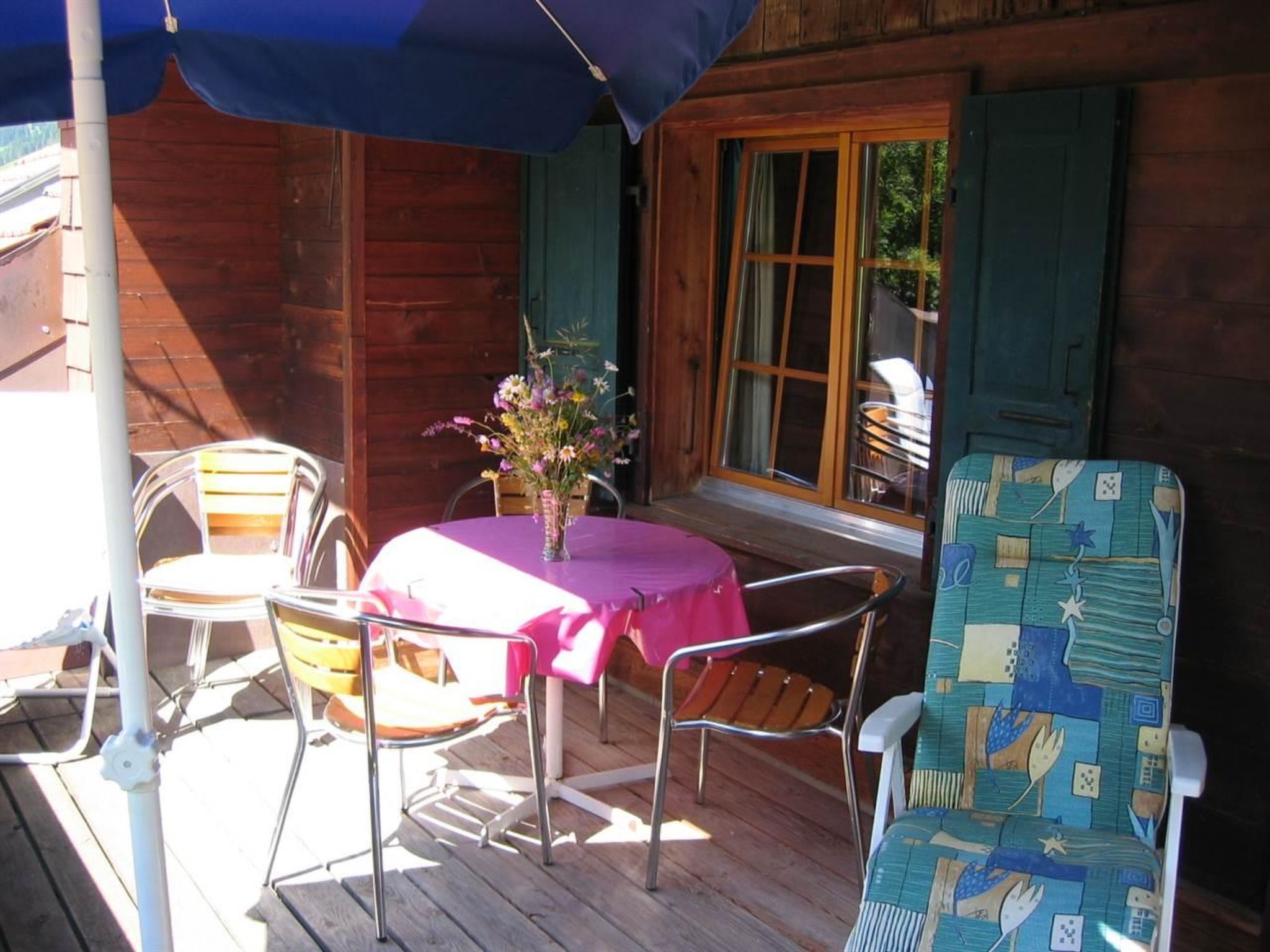 Un patio al aire libre con una mesa y sillas, un jarrón de flores y una cabaña de madera con contraventanas verdes.