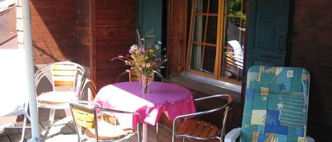 Un patio al aire libre con una mesa y sillas, un jarrón de flores y una cabaña de madera con contraventanas verdes.