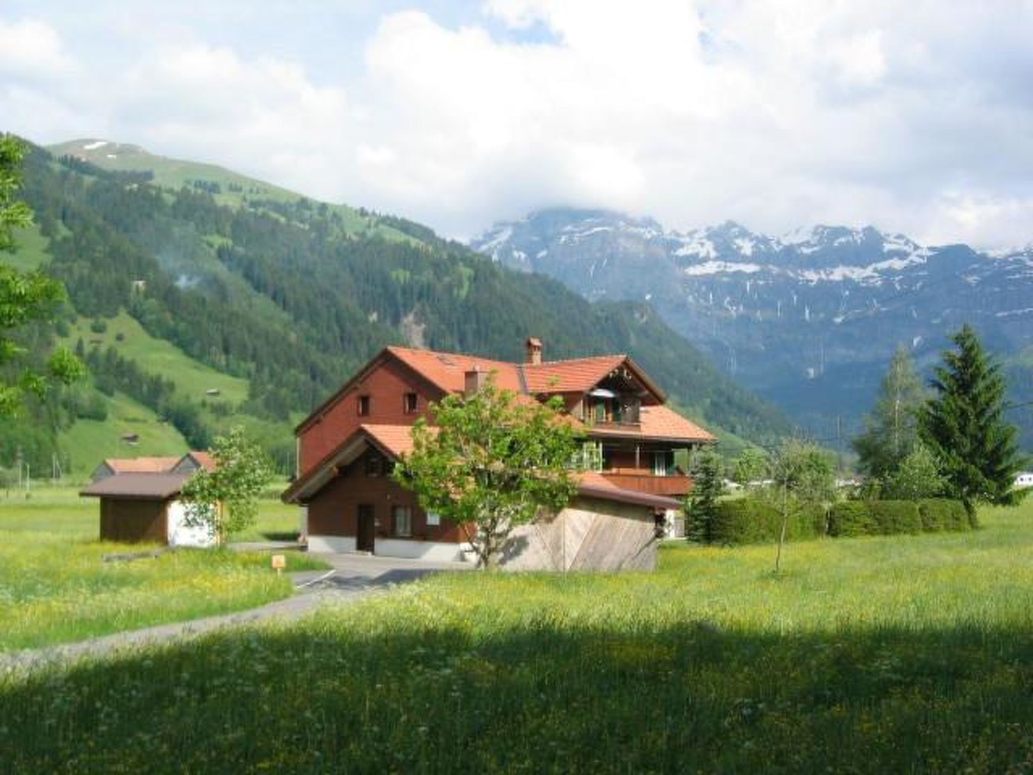 Ein zweigeschossiges Haus mit Balkon, umgeben von einem üppigen Grünland und Bergen im Hintergrund.