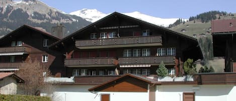 A multi-story chalet with balconies, set against a backdrop of snow-capped mountains.