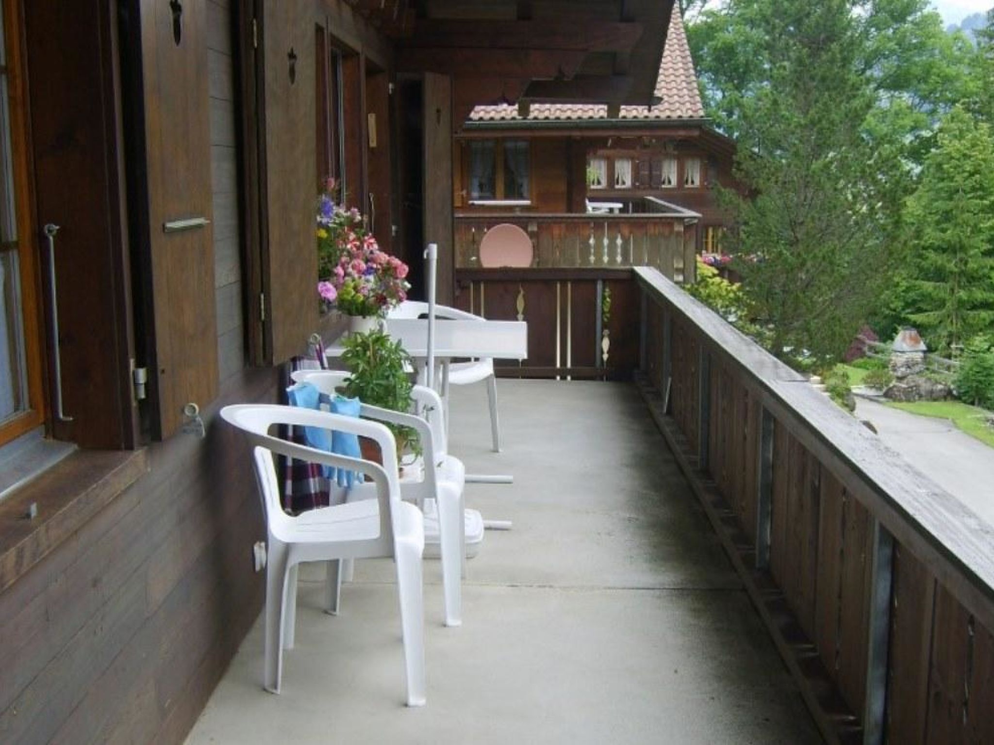 Een houten balkon met witte stoelen en een tafel, met uitzicht op een tuin en een gebouw met een pannendak.