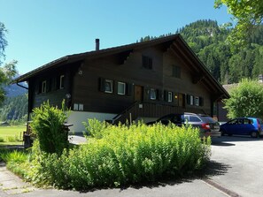 A two-story building with a sloped roof, a staircase leading to the entrance, and a parking area with cars. - Chalet Claudia Appartement/Fewo, Dusche, 1 Schlafraum 2-Bett-Wohnung by Interhome (Lenk)